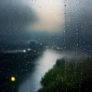 Cristal empañado con gotas de lluvia que distorsionan la vista de un paisaje al fondo, evocando un ambiente melancólico y de incertidumbre, relacionado con el impacto de la DANA.