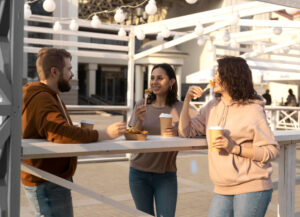 Tres persones joves gaudint d'un àpat informal a l'aire lliure en una terrassa urbana amb estructures blanques i llums penjants. Somriuen i conversen mentre prenen cafè i mengen aperitius.